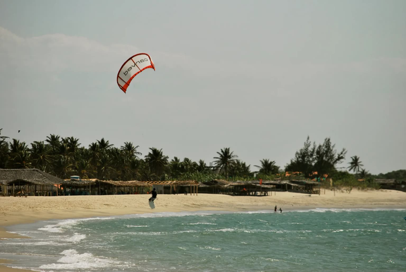 Kitesurfer jumping over the water with the kite in the air, in front of the row of thatched beach bars on Barra Grande beach, Cajueiro da Praia (Piauí), with palm trees behind.