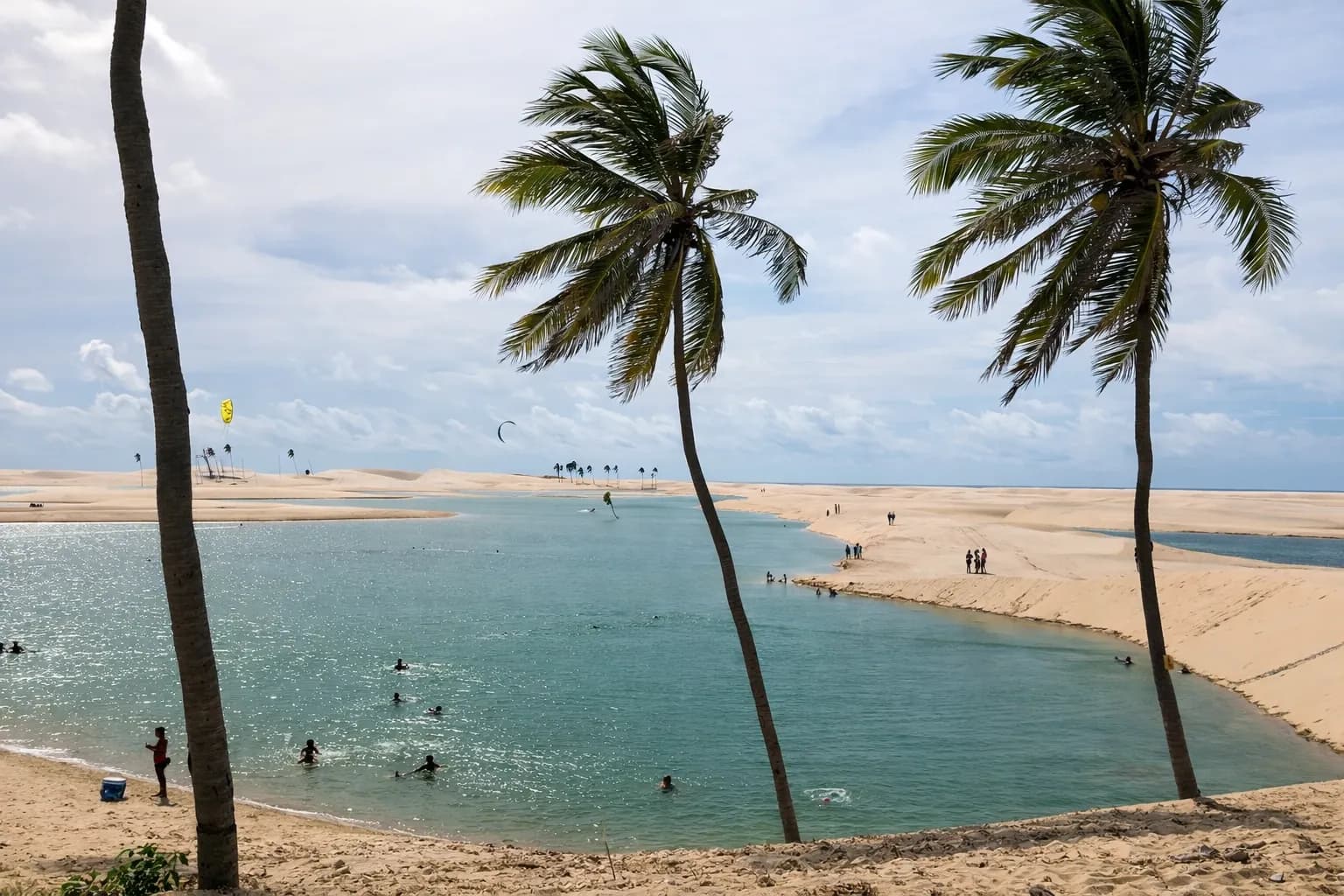 Praia do Arpoador, en Tutóia (MA): agua tranquila entre la playa y un amplio banco de arena, bañistas en la orilla, cocoteros en primer plano y dos kites en el aire sobre las dunas, a lo lejos.